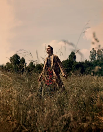 Model wearing a plaid coat walking through tall field grass under an overcast sky, photographed by Takay.