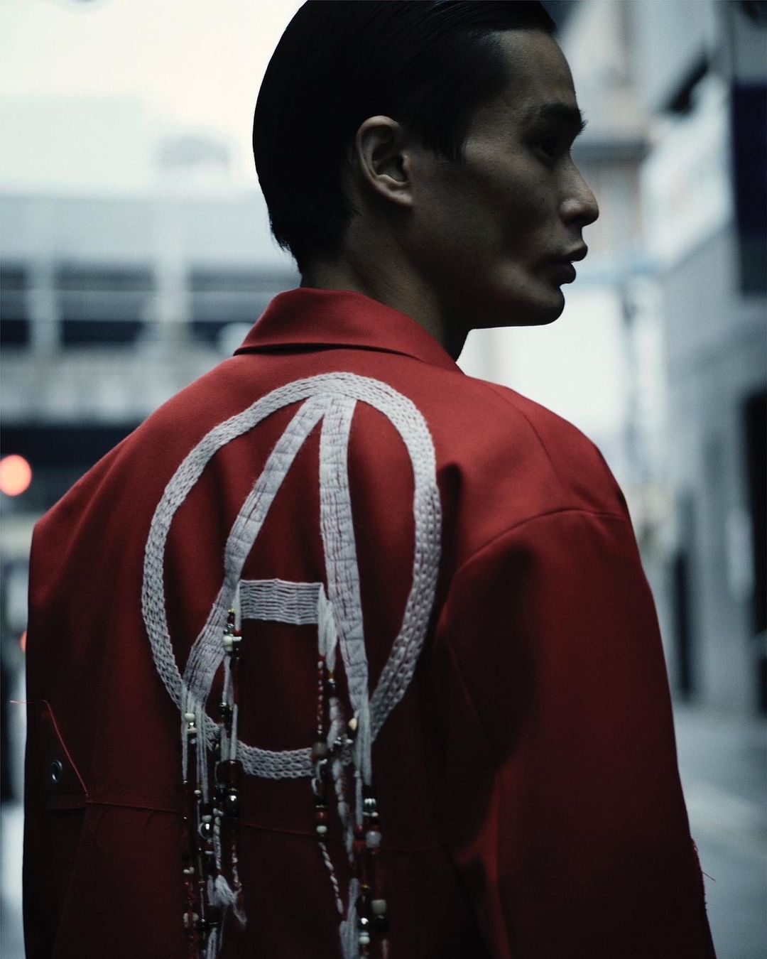 Stylish man in a red jacket with a white circular design stands outdoors in a fashion shot against an urban street backdrop.