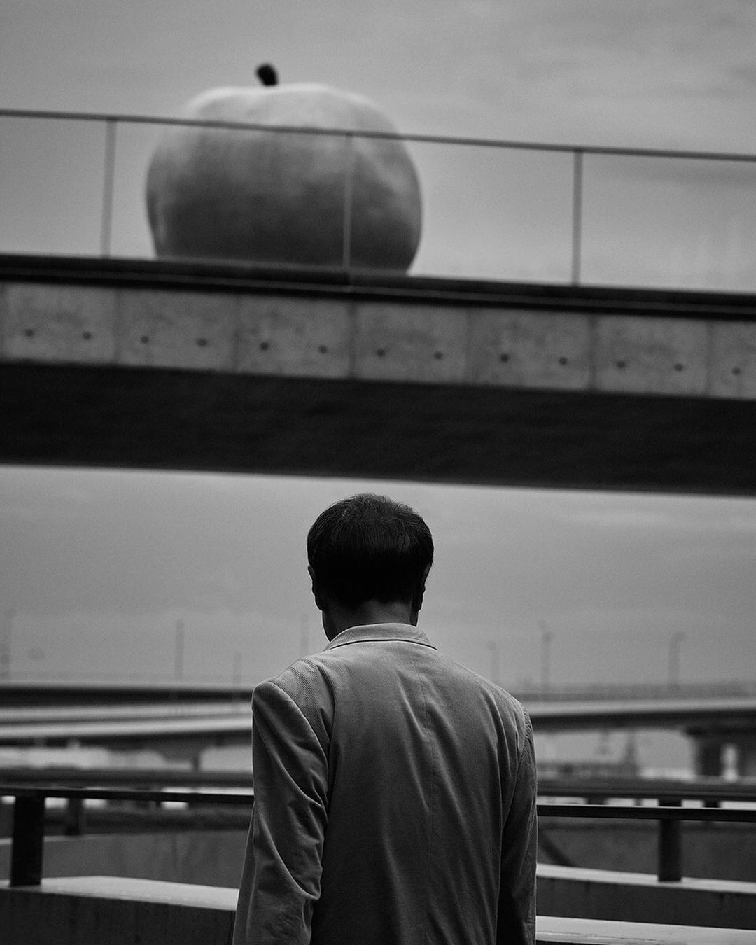 Monochrome photo of a man facing away from the camera, with an oversized apple sculpture on a bridge in the background, black and white.