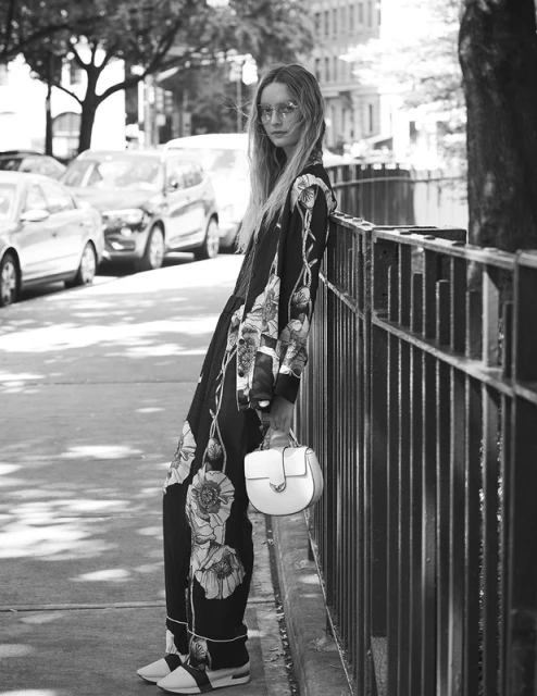 A black-and-white full shot of a woman in a floral pajama-style suit leaning against a metal fence with a white handbag in a city street.