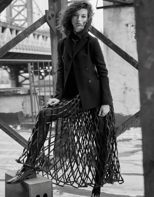 Black and white outdoor fashion portrait of a woman in a blazer and cage skirt, leaning on a metal frame structure with a building backdrop.