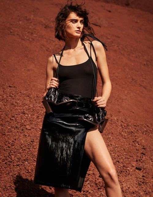 A brunette woman in a black top and skirt poses against a textured red background, showcasing an edgy and modern fashion aesthetic.