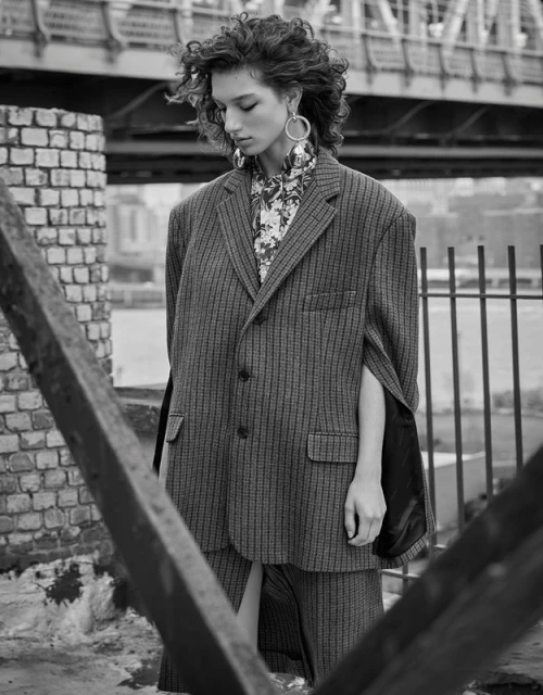 A young woman with curly hair poses wearing an oversized pinstripe blazer and skirt with a floral shirt in an urban setting.