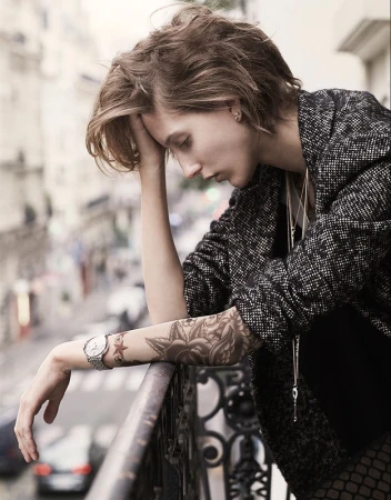 A woman with short brown hair leaning on a balcony railing, wearing a textured tweed jacket and tattoos on her forearm, photographed by Takay.