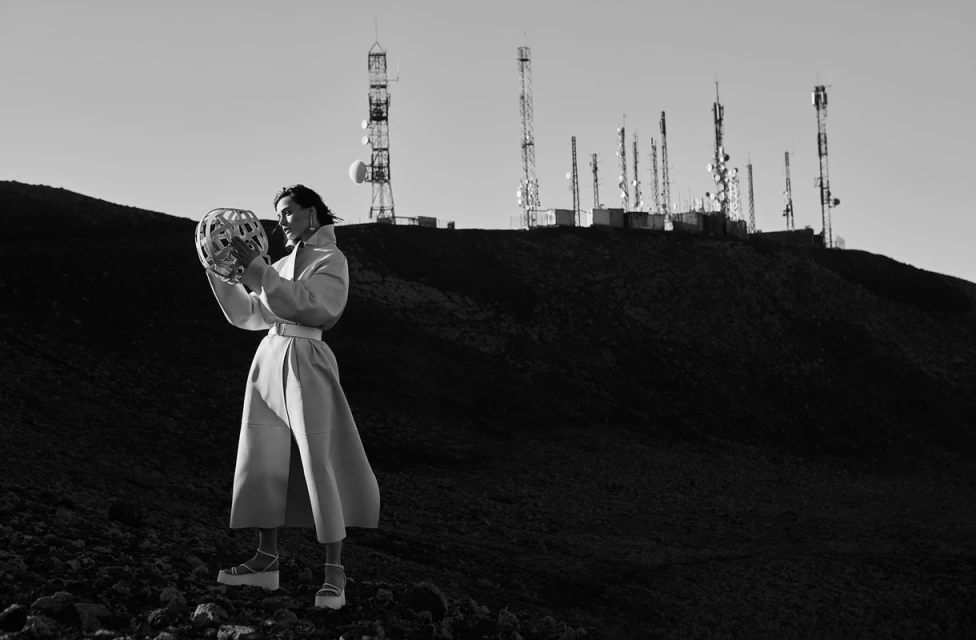 A woman in a coat poses on a dark hillside with a decorative sphere, while radio communication towers populate the distant mountain top.