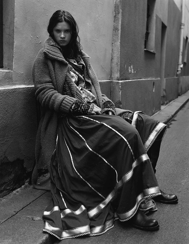 Monochrome portrait of a young woman in a coat and long skirt sitting on a street, showcasing fashion and urban style.