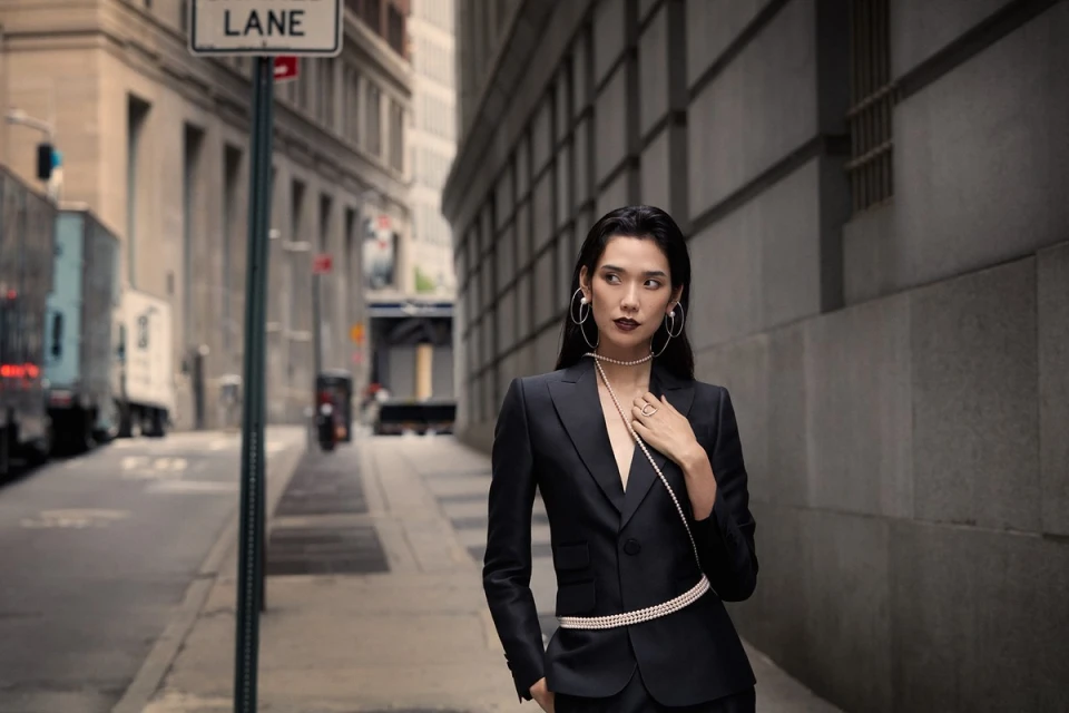 Elegant Asian woman in a black suit poses on a city street. She wears pearl jewelry and hoop earrings, with a blurred cityscape backdrop.