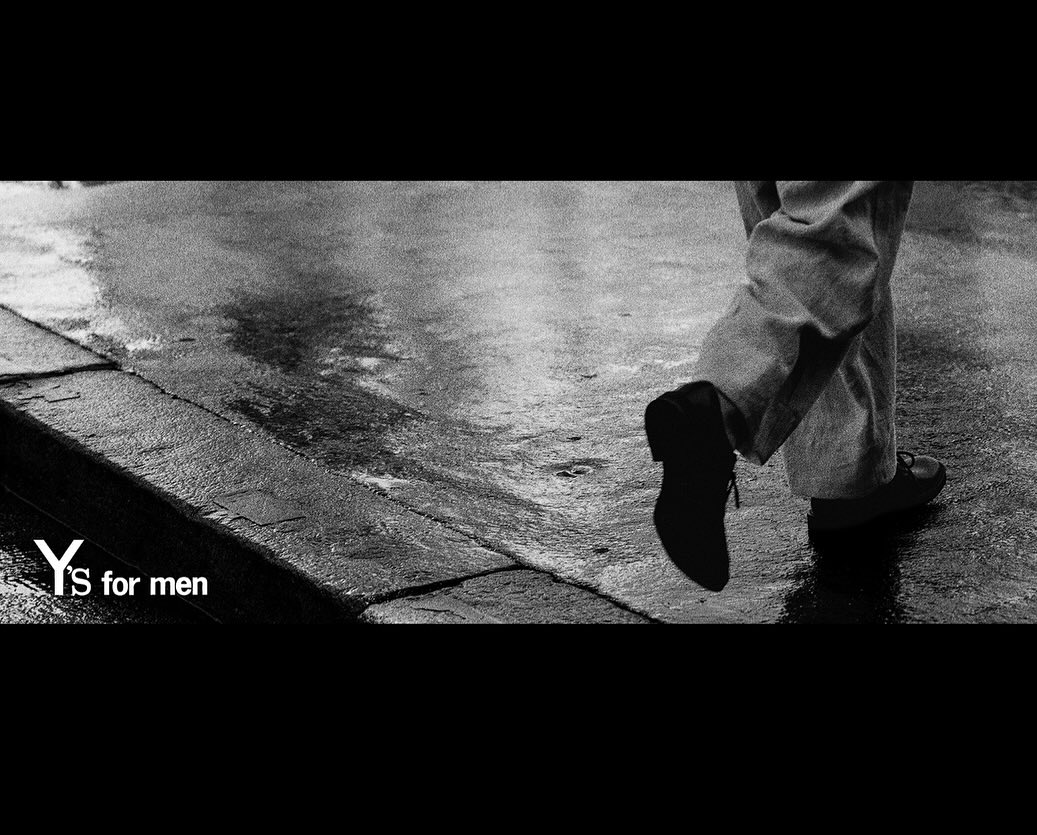 Monochrome fashion shot of a person walking on a wet street, wearing dress pants and shoes; logo, "Y'S for men", at the bottom left.