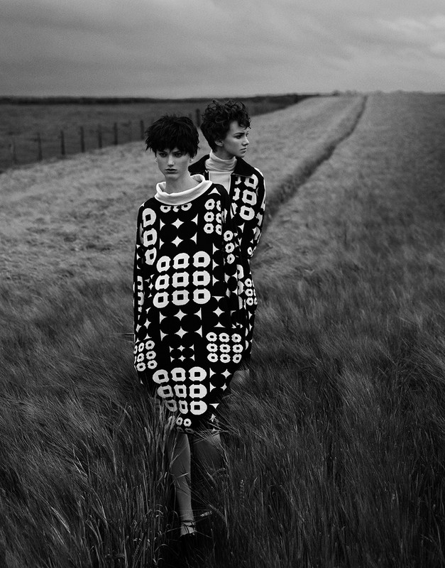 Black-and-white outdoor fashion shot of two women standing in tall grass, wearing patterned dresses. Moody sky.