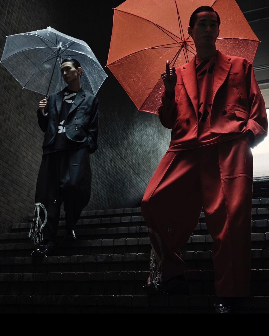 Two Asian men in stylish suits descend concrete stairs under umbrellas, one giving the middle finger in a darkly lit, urban scene.