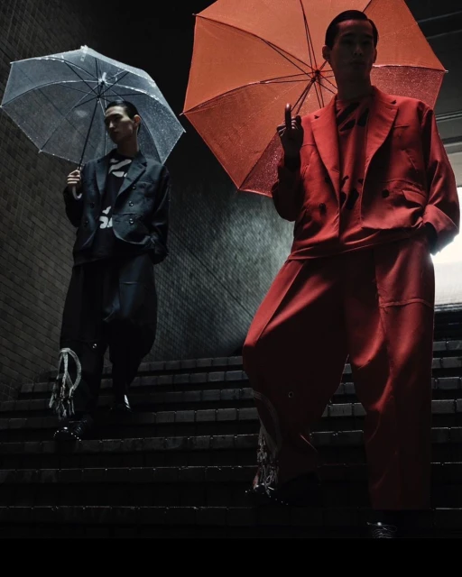 Two Asian men in stylish suits descend concrete stairs under umbrellas, one giving the middle finger in a darkly lit, urban scene.