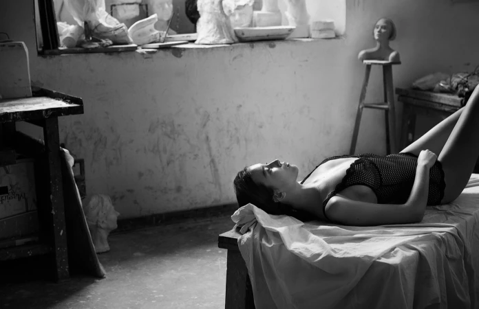 Woman in dark lace bodysuit lying on a draped workbench inside an artist studio with plaster busts in the background