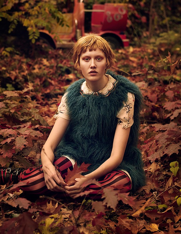 Young female model with reddish-blonde hair, wearing a green fur vest, is sitting in a forest surrounded by fallen autumn leaves.