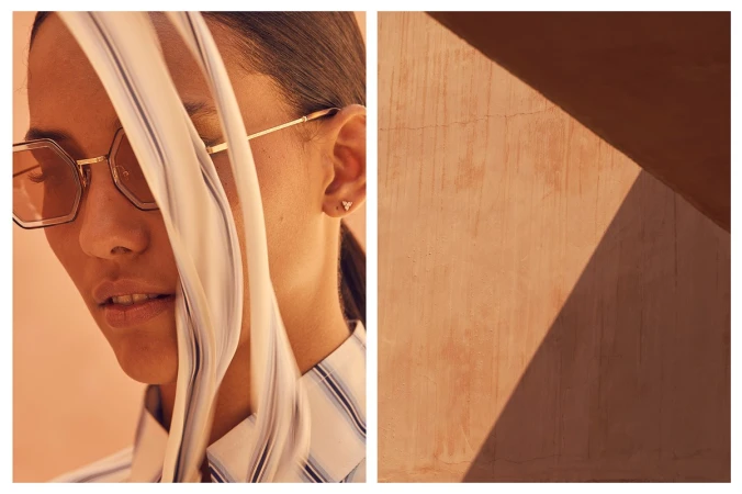 Close-up portrait of a woman wearing geometric sunglasses and a striped shirt, framed beside a sunlit wall panel.