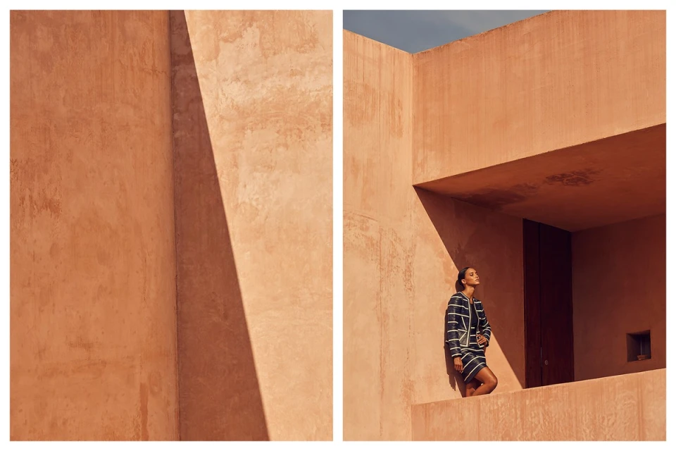 Fashionable woman in striped suit posing against a peach building, bathed in sunlight, highlighting architectural lines and shadows.