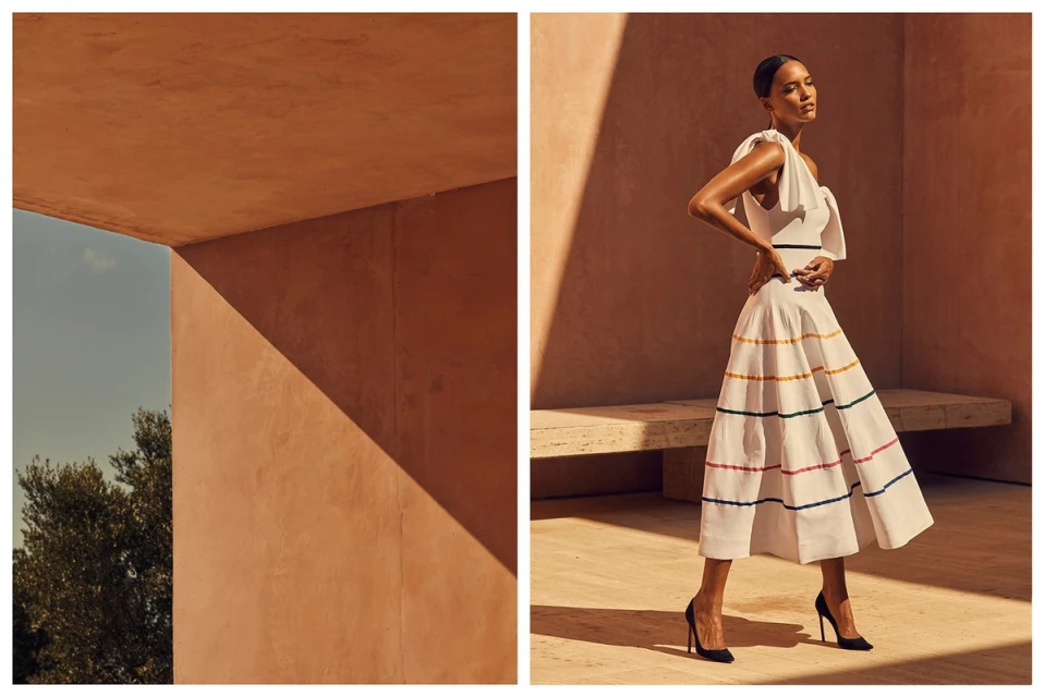 Woman in white sundress posing by architectural building in sun. Fashion editorial image featuring a model in front of modern architecture.