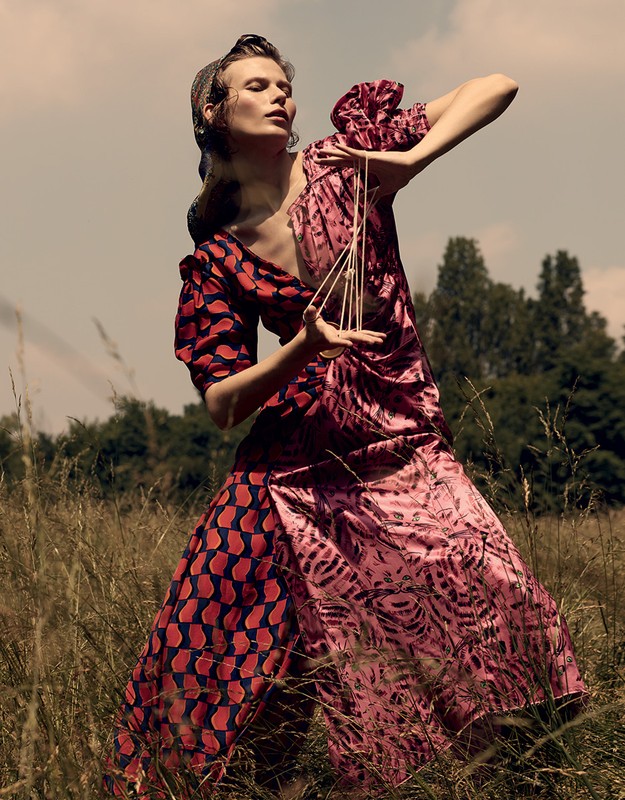Woman in patterned dress holds geometric sculpture in a grassy field with trees and blue sky; fashion editorial shot, outdoors.