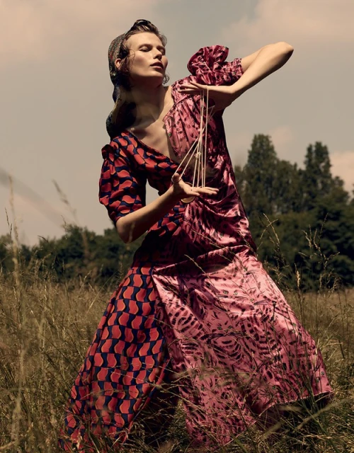 Woman in patterned dress holds geometric sculpture in a grassy field with trees and blue sky; fashion editorial shot, outdoors.