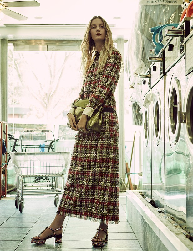 A stylish woman in a plaid skirt suit poses inside a bright laundromat, holding a gold bag, with washing machines in the background.