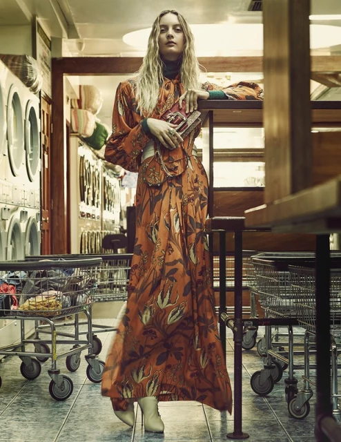 A woman with blonde hair poses in a laundromat wearing an orange floral dress and white boots, standing near laundry carts.