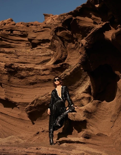 A woman in a black sequined dress poses in a desert landscape with rocky formations, wearing sunglasses and black boots against a blue sky.