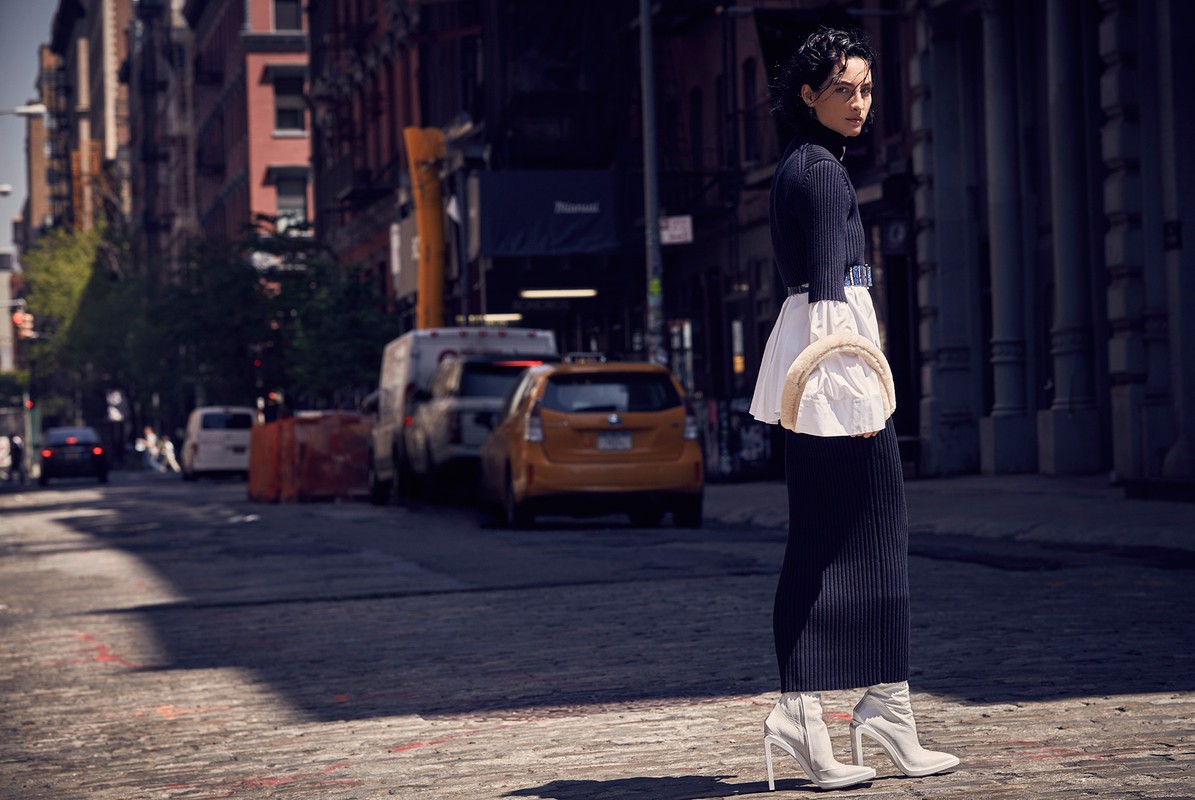 Woman in a dark turtleneck dress, white boots, and a matching bag standing on a brick street in an urban setting with buildings and cars.
