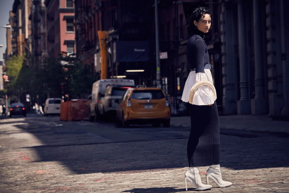 Woman in a dark turtleneck dress, white boots, and a matching bag standing on a brick street in an urban setting with buildings and cars.