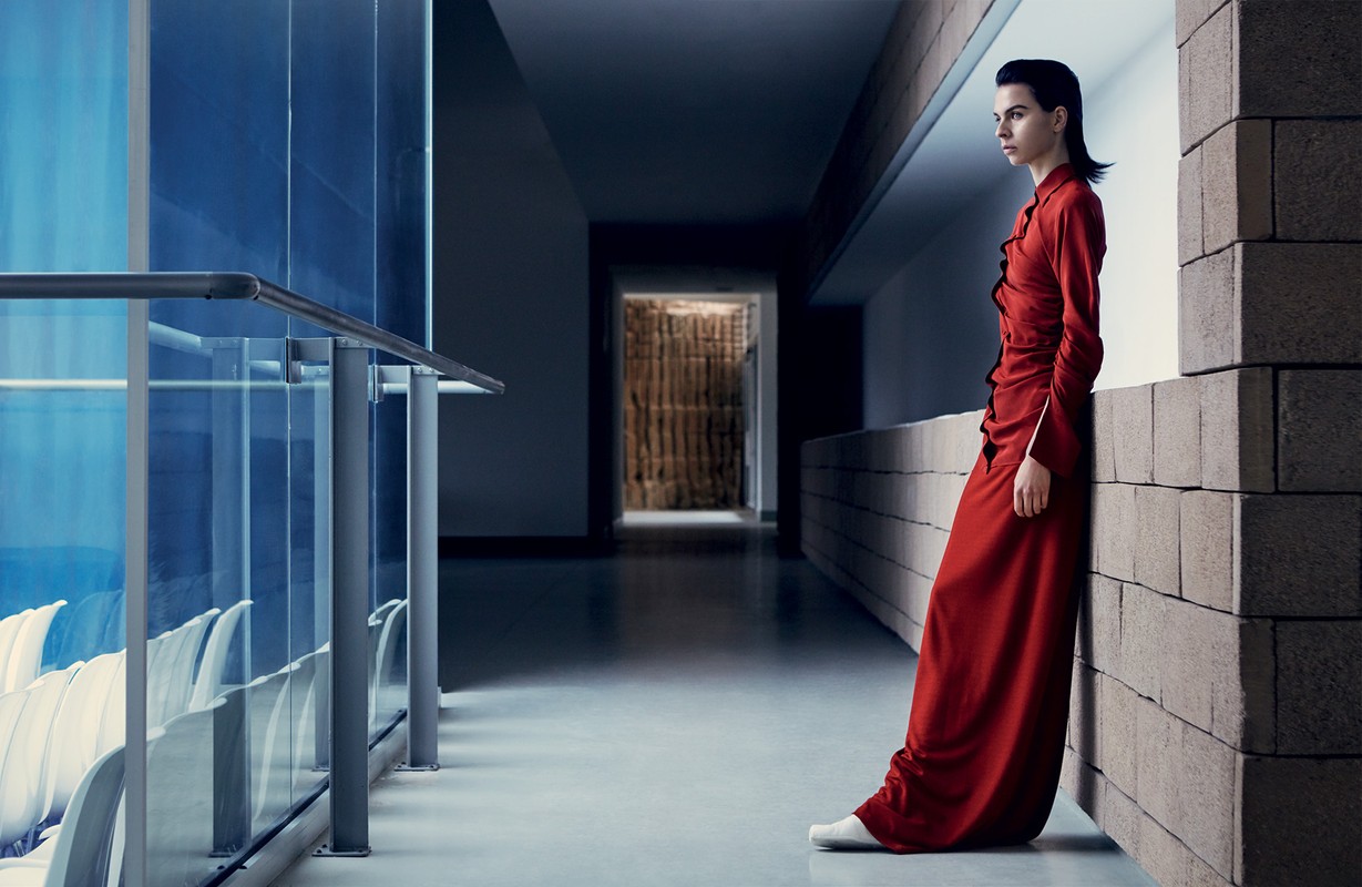 A woman in a red dress leans against a brick wall in a long hallway with cool lighting and modern design elements, creating an editorial fashion image.