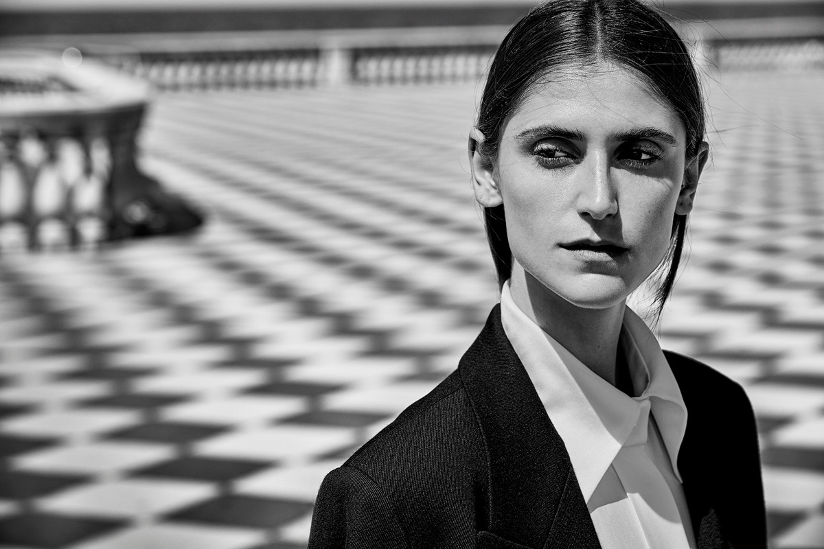 Monochrome portrait of a brunette woman in a blazer and white shirt, set against a backdrop of patterned floor tiles and sunlight.
