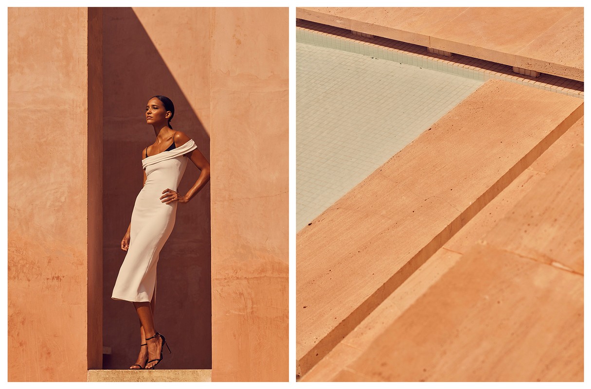 Elegant woman in white off-the-shoulder dress and black heels poses against a textured orange wall with natural sunlight and shadows.
