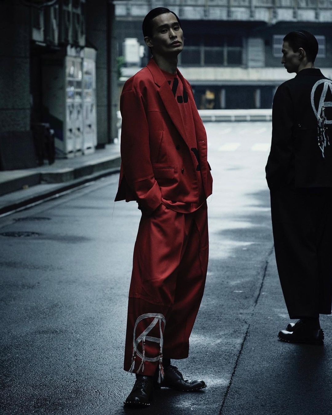 Two male fashion models pose on a city street with buildings. One model wears a red suit and the other wears black clothing.