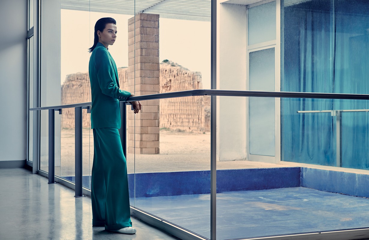 A woman in a green suit stands by a window with an outdoor view of a textured stone structure, bathed in natural light.