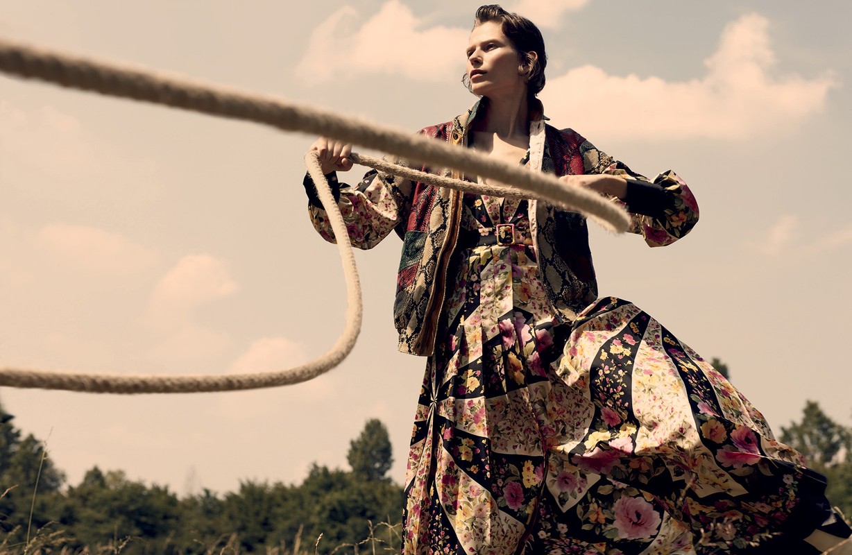 Woman in a floral dress and jacket holds a rope in a sunlit field, with trees and a blue sky visible in the background.