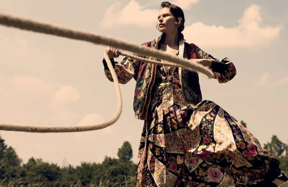 Woman in a floral dress and jacket holds a rope in a sunlit field, with trees and a blue sky visible in the background.
