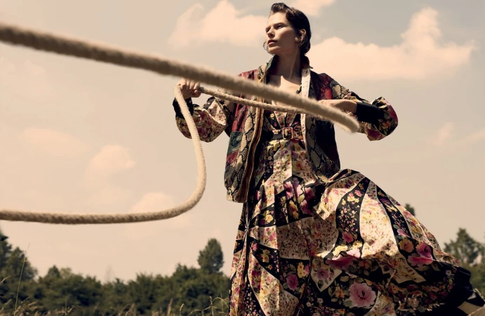 Woman in a floral dress and jacket holds a rope in a sunlit field, with trees and a blue sky visible in the background.