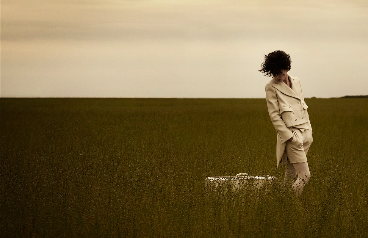 Adult in a beige suit standing in a field with tall grass, next to a matching suitcase, under a cloudy sky.