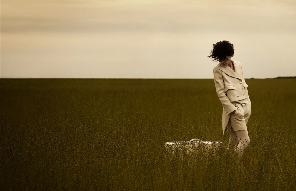 Adult in a beige suit standing in a field with tall grass, next to a matching suitcase, under a cloudy sky.