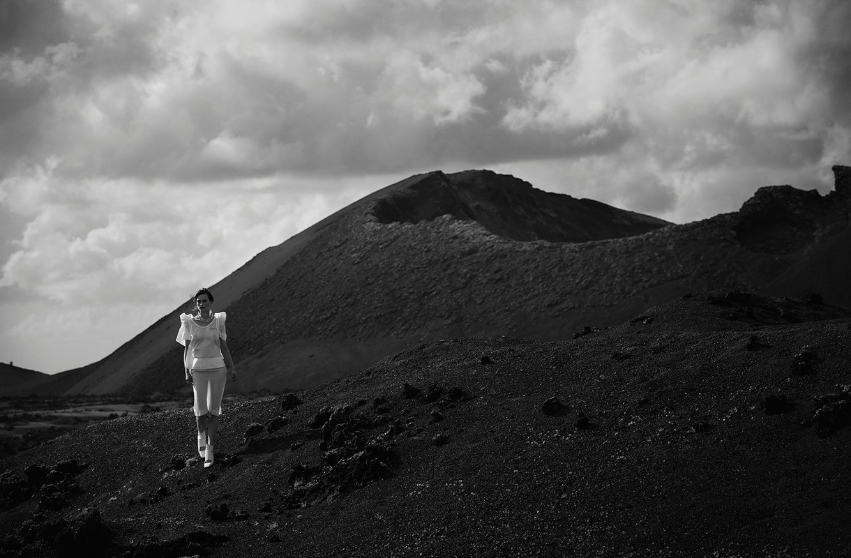 Woman in white outfit walks on a black volcanic landscape beneath a cloudy sky, with a volcanic crater in the background in black and white.