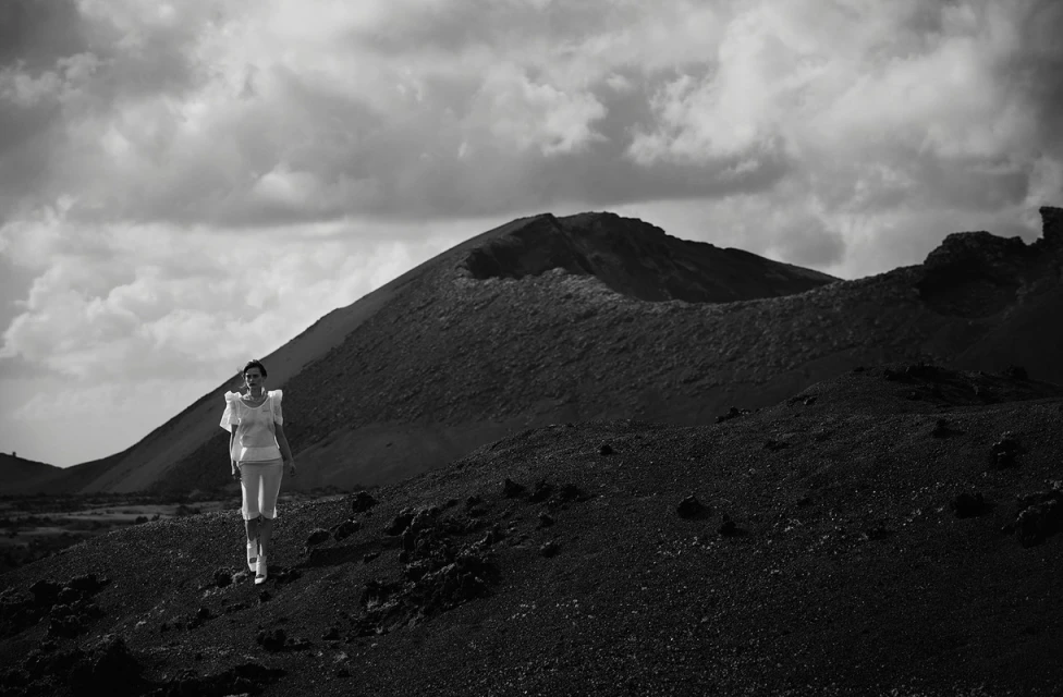 Woman in white outfit walks on a black volcanic landscape beneath a cloudy sky, with a volcanic crater in the background in black and white.