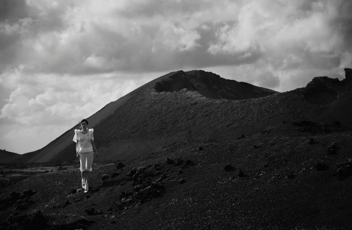 Woman in white outfit walks on a black volcanic landscape beneath a cloudy sky, with a volcanic crater in the background in black and white.