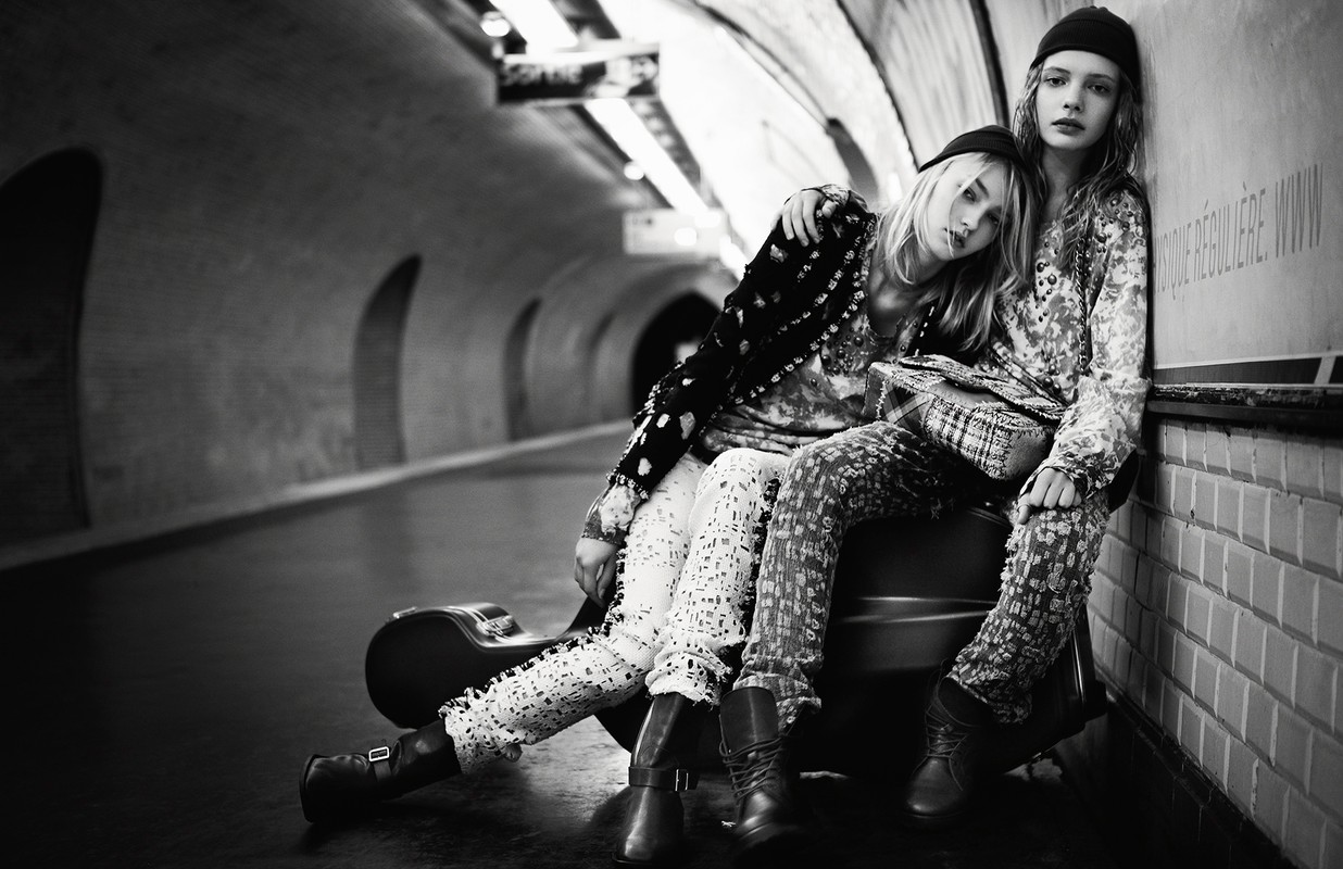 Two young women in patterned outfits sit in a subway tunnel with a guitar case, looking directly at the camera in a black-and-white shot.