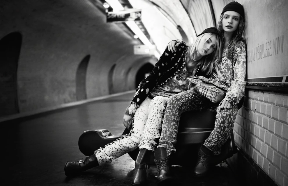 Two young women in patterned outfits sit in a subway tunnel with a guitar case, looking directly at the camera in a black-and-white shot.