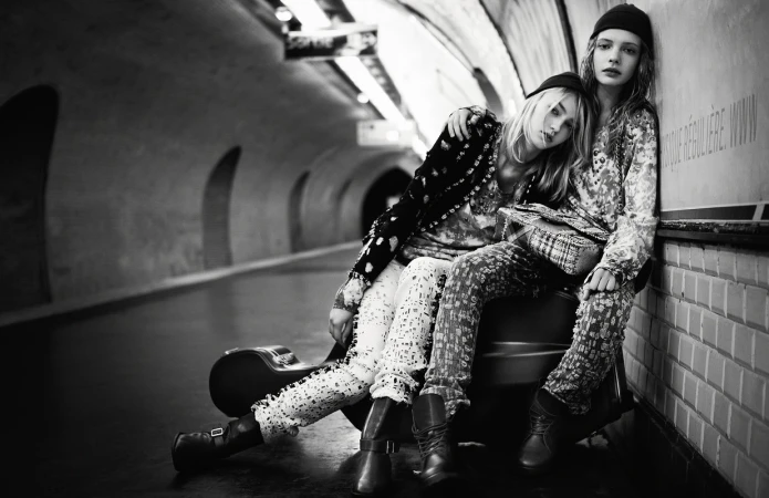 Two young women in patterned outfits sit in a subway tunnel with a guitar case, looking directly at the camera in a black-and-white shot.
