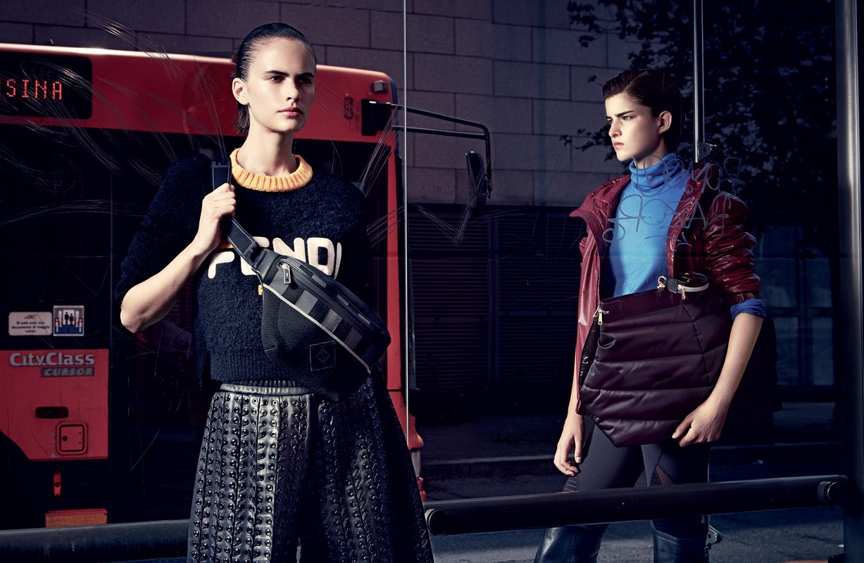 Two fashion models pose in front of a bus with a bag. One model is wearing a Fendi sweater, the other is wearing a blue turtleneck and jacket.