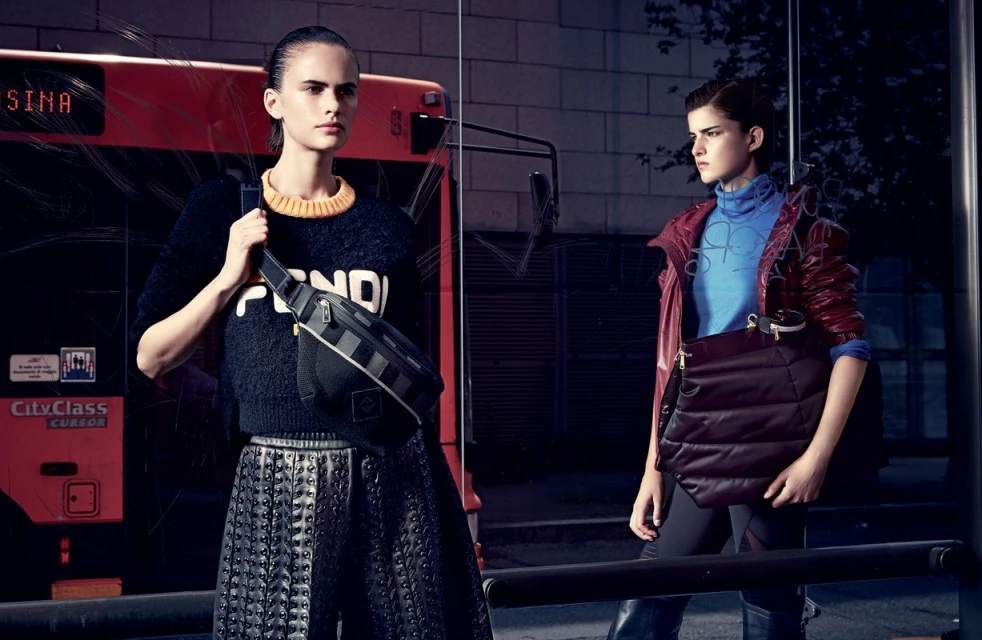 Two fashion models pose in front of a bus with a bag. One model is wearing a Fendi sweater, the other is wearing a blue turtleneck and jacket.