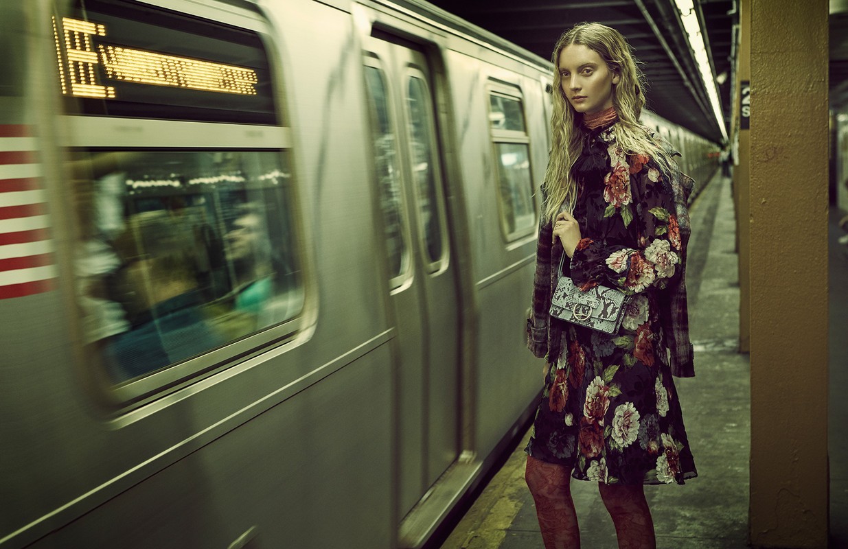 A woman with long blonde hair stands on a subway platform next to a moving train, wearing a floral dress and holding a small bag.
