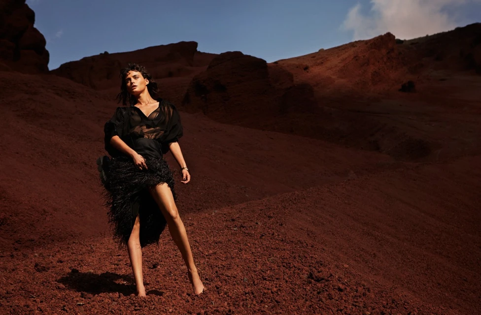 A model in a black sheer dress poses on a red clay hillside with a blue sky in the background, fashion editorial, outdoors.