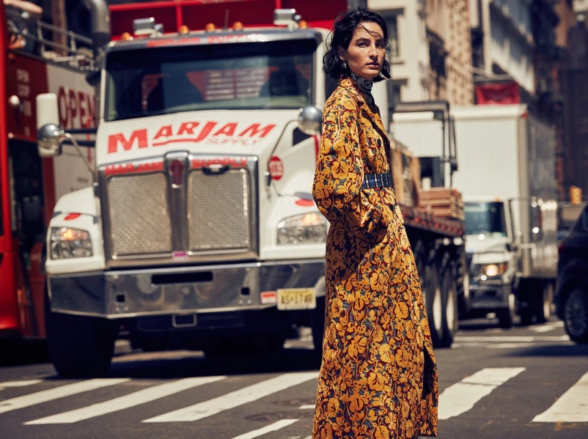 A brunette woman in a floral trench coat stands on a crosswalk in an urban setting with a large commercial truck in the background.