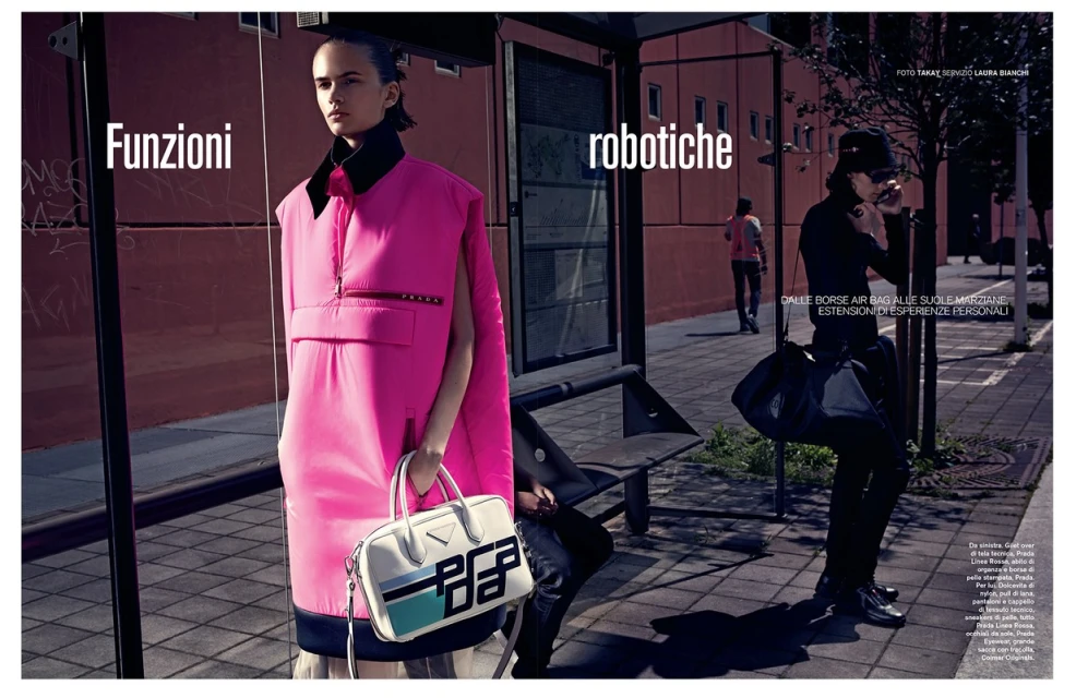 Editorial fashion shot at a bus stop: a woman in a pink sleeveless dress holds a Prada handbag, while another waits in the background.