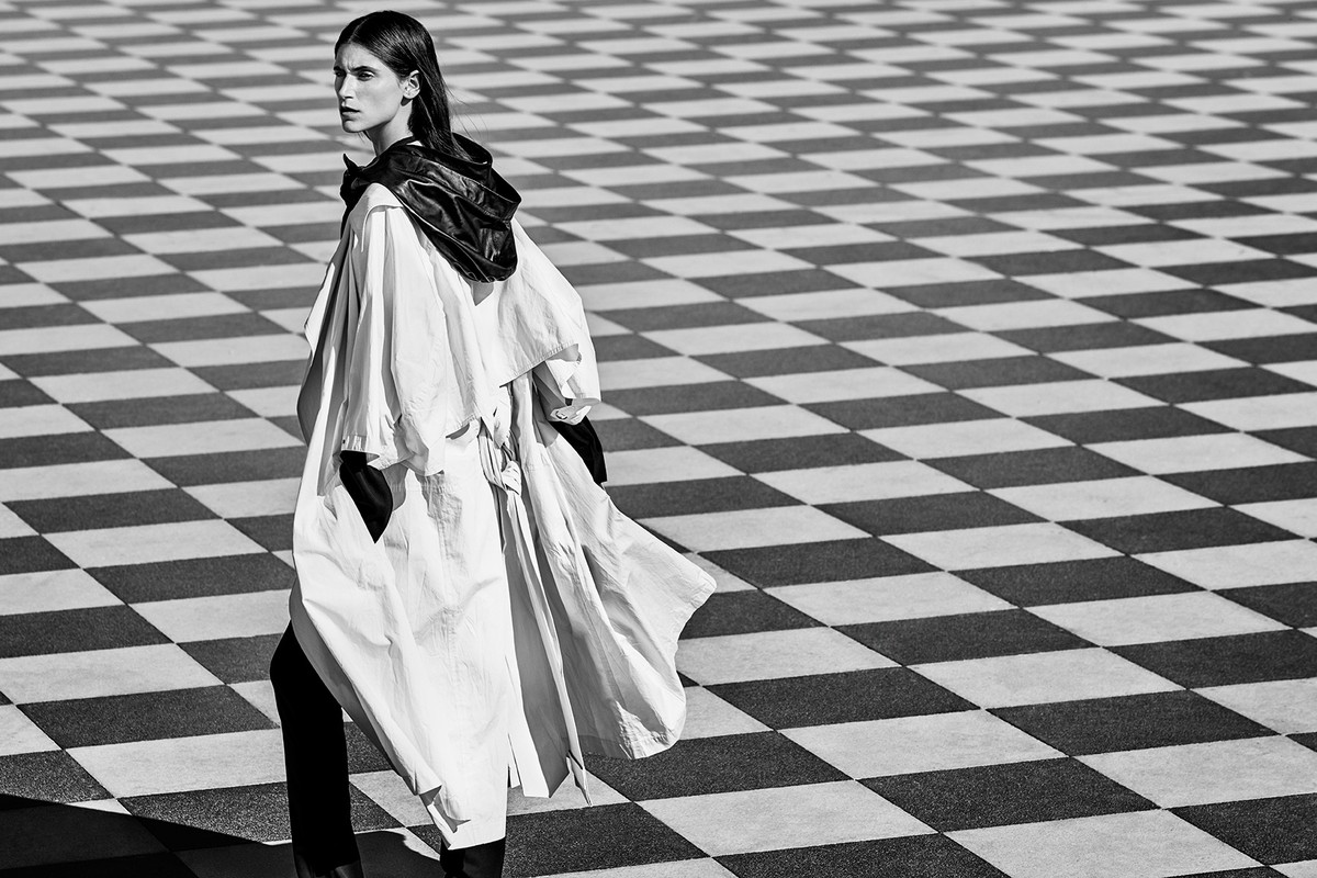 An adult woman in black pants and light coat walks across a checkered surface, looking left in a black and white outdoor shot.